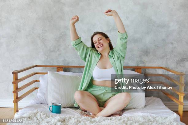 pretty woman sit on bed stretched her arms, makes exercise while awakened with cup of morning coffee - mature woman beauty arm stockfoto's en -beelden