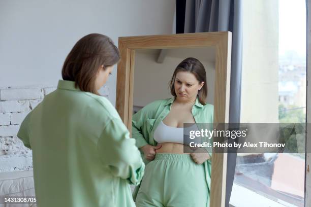 size plus woman looking by reflection in mirror feels frustrated and dissatisfied by overweight - dicke frau stock-fotos und bilder