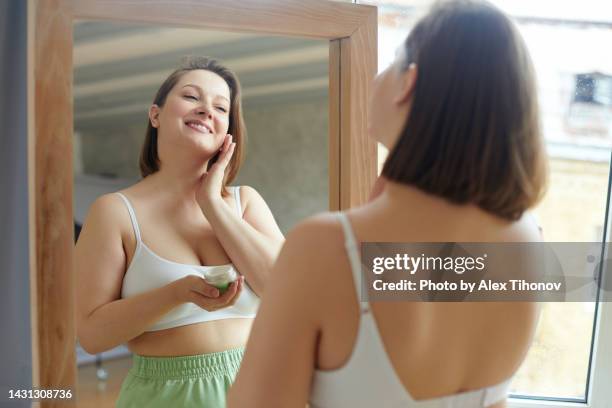 rear view of young plus size adult woman apply facial cream reflecting in mirror do morning routine at home - perfect female body shape fotografías e imágenes de stock