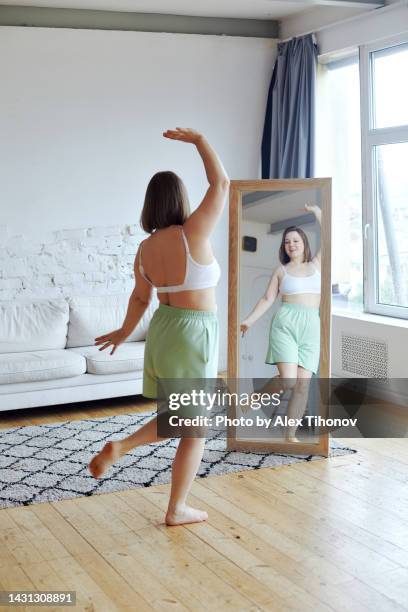 vertical image rear view plus size woman dancing standing barefoot in living room in front of mirror - fat woman dancing stockfoto's en -beelden