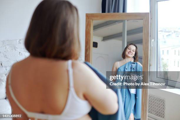 smiling oversize woman reflecting in the mirror, choose apparel to wear in the morning - woman jacket stock pictures, royalty-free photos & images