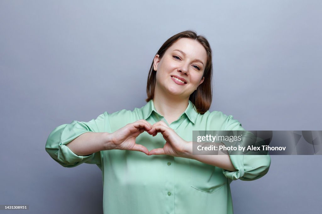 Headshot portrait of loving young female posing on grey studio wall background smile look at camera showing with joined fingers heart symbol, makes like gesture, express gratitude and appreciation