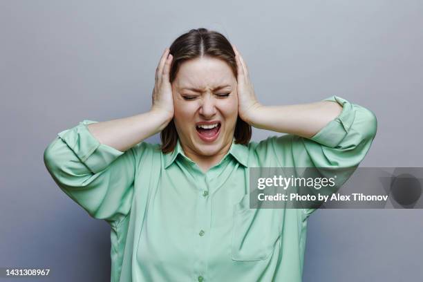 woman screaming with eyes closed on grey studio background, headshot portrait - hands covering ears stock-fotos und bilder