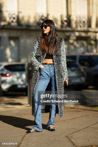 Leia Sfez wears black sunglasses, a black cropped top, a gray and black fluffy long coat, a black shiny leather belt, blue denim large pants, black...