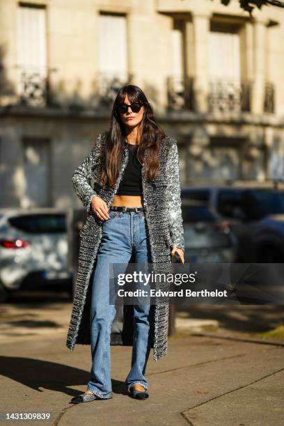 Leia Sfez wears black sunglasses, a black cropped top, a gray and black fluffy long coat, a black shiny leather belt, blue denim large pants, black...