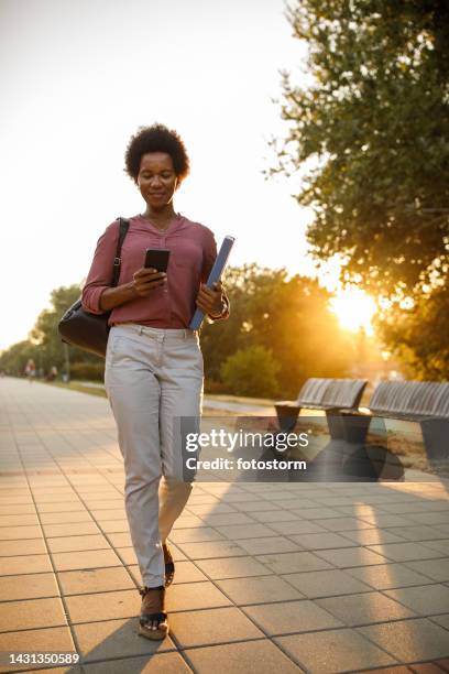 businesswoman walking down the sunny city street and using smart phone when going to work - golden hour woman stock pictures, royalty-free photos & images