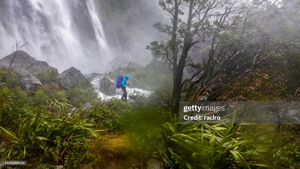 Woman backpacker hiking in front of raging Earland Falls, Routeburn Track, Fiordland National Park, New Zealand.