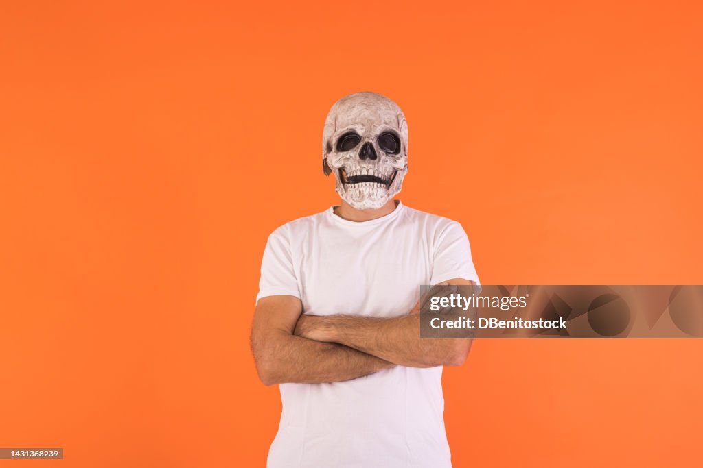 Man wearing skull mask and white t-shirt, with his arms crossed, celebrating Halloween, on an orange background. Concept of celebration, day of the dead, all saints' day, dressing up and carnival.