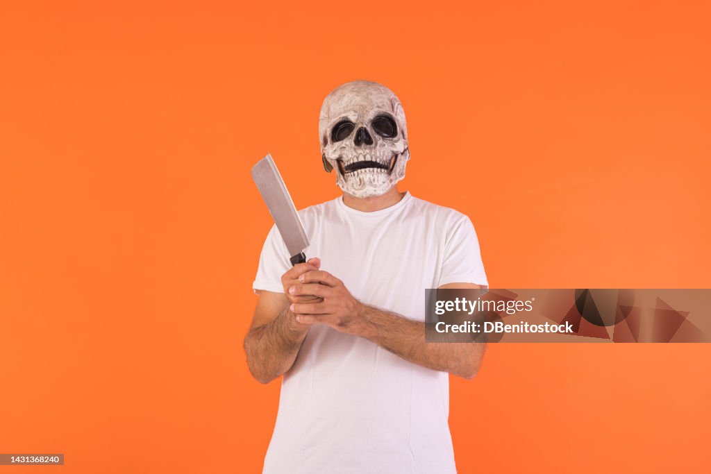 Man wearing skull mask and white t-shirt, holding a knife, celebrating Halloween, on an orange background. Concept of celebration, day of the dead, all saints' day, dressing up and carnival.