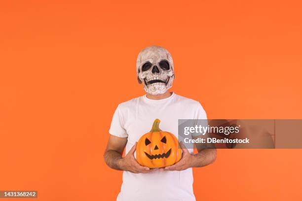 man wearing skull mask and white t-shirt, holding a pumpkin, with copy space, celebrating halloween, on an orange background. concept of celebration, day of the dead, all saints' day and carnival. - cover monster face stock pictures, royalty-free photos & images