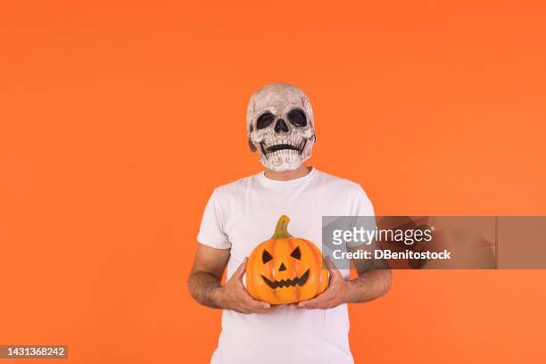 man wearing skull mask and white t-shirt, holding a pumpkin, with copy space, celebrating halloween, on an orange background. concept of celebration, day of the dead, all saints' day and carnival. - cover monster face bildbanksfoton och bilder