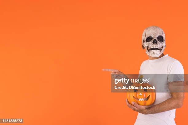 man wearing skull mask and white t-shirt, holding a pumpkin and pointing to the side with his finger, with copy space, celebrating halloween, on an orange background. concept of celebration, day of the dead. - cover monster face bildbanksfoton och bilder