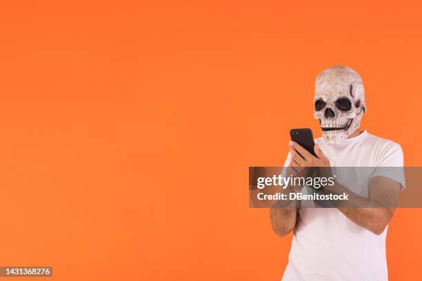 man with skull mask and white t-shirt, consulting his mobile phone, celebrating halloween, on an orange background. concept of celebration, day of the dead, all saints day and carnival. - cover monster face stock pictures, royalty-free photos & images