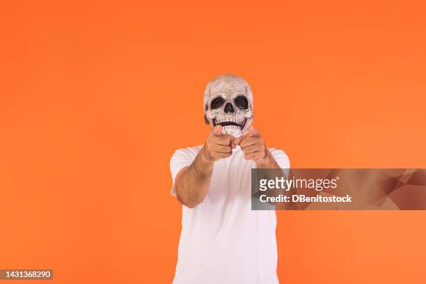 man with skull mask and white t-shirt, pointing fingers at camera, celebrating halloween, on orange background. concept of celebration, day of the dead, all saints day and carnival. - all shirts stock-fotos und bilder