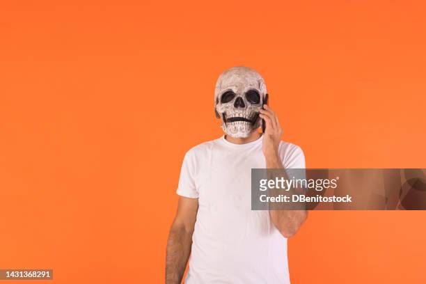 man with skull mask and white t-shirt, talking on his mobile phone, celebrating halloween, on an orange background. concept of celebration, day of the dead, all saints day and carnival. - cover monster face stock pictures, royalty-free photos & images