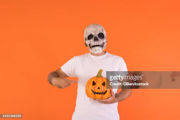 man wearing skull mask and white t-shirt, pointing at a pumpkin, with copy space, celebrating halloween, on an orange background. concept of celebration, day of the dead, all saints' day,and carnival. - cover monster face bildbanksfoton och bilder