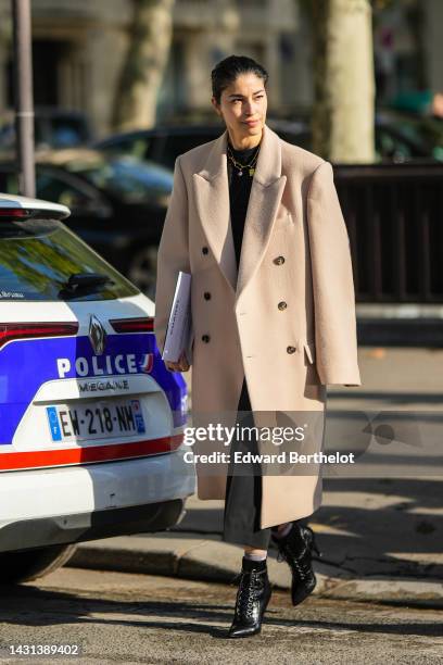 Caroline Issa wears a gold large chain pendants necklace, a black pullover, a beige wool oversized long coat, black suit pants, black shiny varnished...