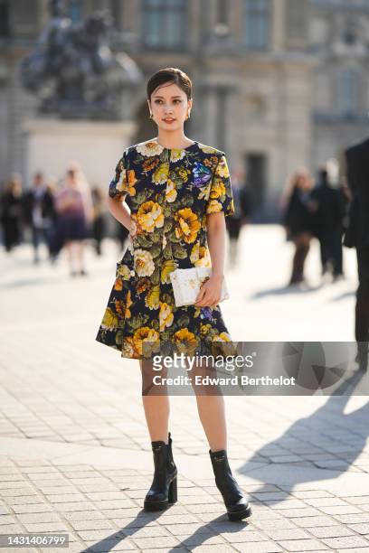 Guest wears gold earrings, a black with yellow / green flower print pattern short sleeves / knees dress, a white shiny leather Petite Malle handbag...