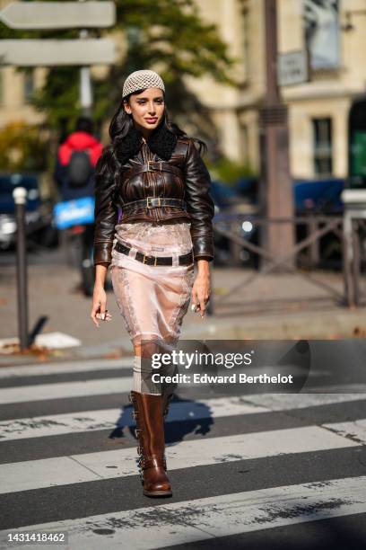 Guest wears a beige mesh / fishnet beanie, a brown shiny leather belted jacket, a black shiny leather belt, gold and diamond rings, a beige silk with...