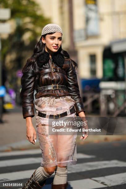 Guest wears a beige mesh / fishnet beanie, a brown shiny leather belted jacket, a black shiny leather belt, gold and diamond rings, a beige silk with...