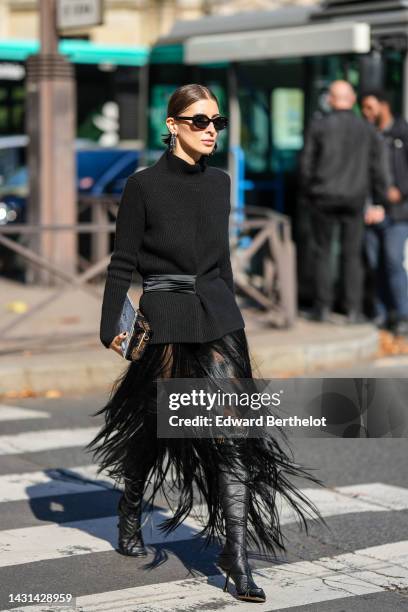 Guest wears black sunglasses, black shiny leather nailed / studded earrings, a black ribbed wool turtleneck belted jacket, a black feathers long...