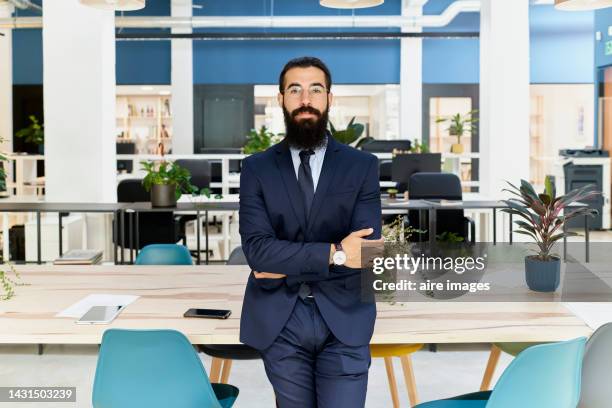 man dressed in a suit with glasses and a beard standing leaning on the desk with his arms crossed denoting authority facing the camera is inside his workplace. - business person facing away from camera stockfoto's en -beelden