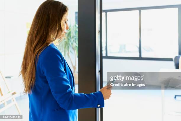 young woman with long hair standing in profile inside her workplace, serious and worried, checking her cell phone, through the window the sunlight is coming in. - coming back stock pictures, royalty-free photos & images