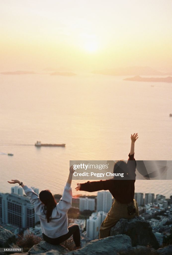 Two young girls are sitting on the mountain in front of the sunset, raising their hands.
