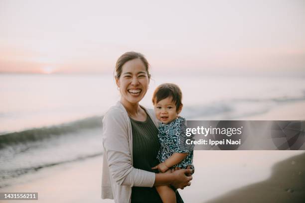 happy mother holding baby on beach at sunset - familia de dos generaciones fotografías e imágenes de stock