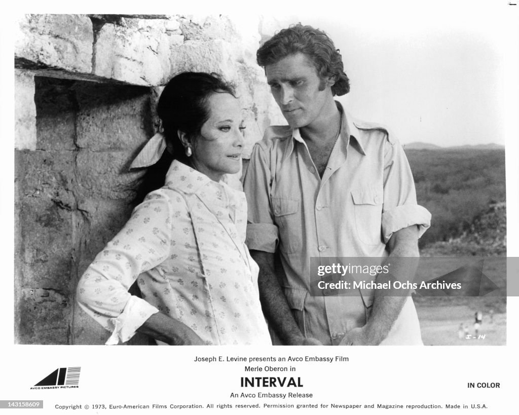 Merle Oberon leaning against a wall as Robert Wolders stands looking