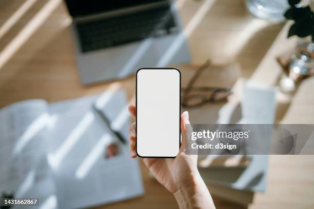 cropped hand of woman holding up a smartphone against wooden office desk, with laptop, books, eye glasses and a glass of water on the table. flat lay shot. top view workspace. business and communication concept - office work flat lay stock pictures, royalty-free photos & images