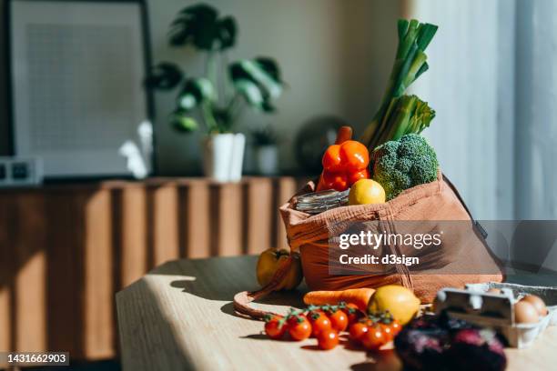 eco-friendly reusable shopping bag with multi-coloured fresh vegetables and groceries on the table at home. shopping with reusable grocery bag for plastic free and waste-free life. responsible shopping. zero waste and sustainable lifestyle concept - groceries stock-fotos und bilder