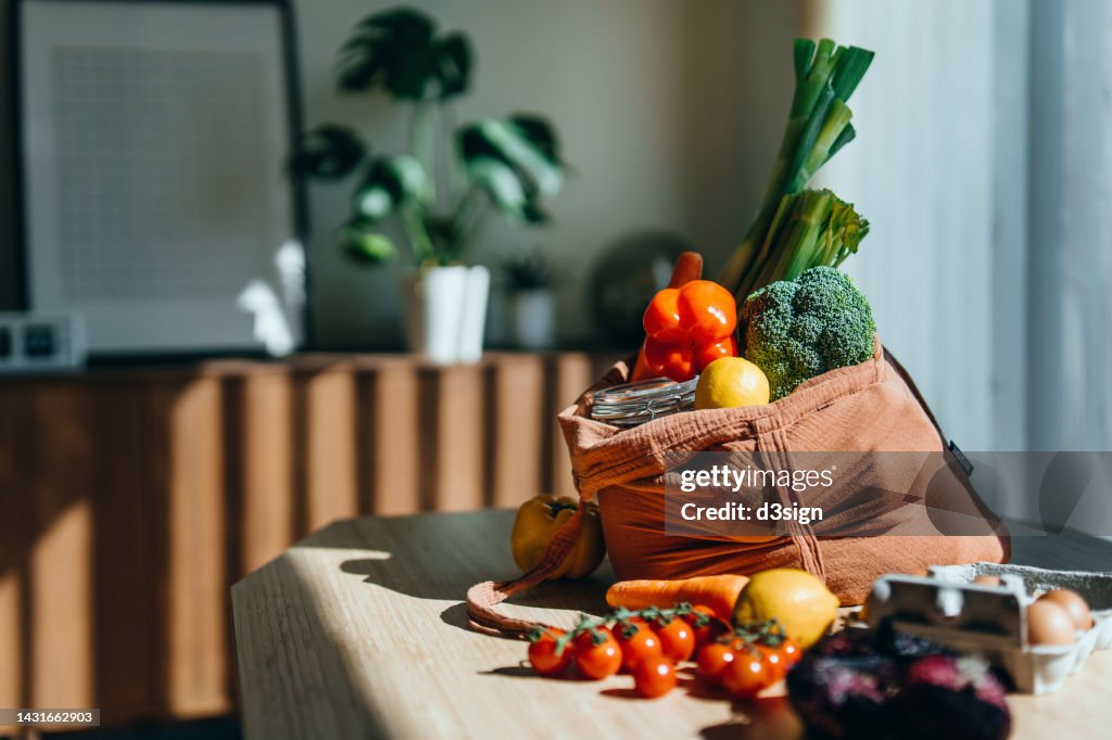 Eco-friendly reusable shopping bag with multi-coloured fresh vegetables and groceries on the table at home. Shopping with reusable grocery bag for plastic free and waste-free life. Responsible shopping. Zero waste and sustainable lifestyle concept