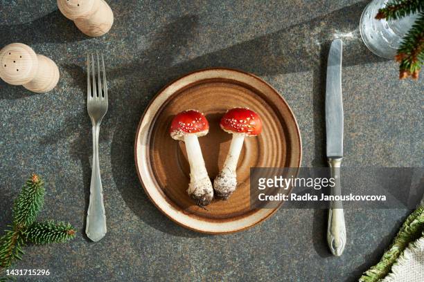 two red fly agaric or amanita mushrooms on a plate on a table with fork and knife - poisonous mushroom fotografías e imágenes de stock