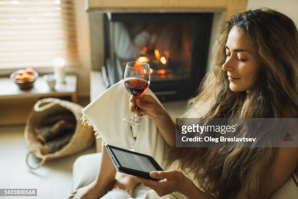 woman reading ebook on digital tablet and drinking red wine. relaxing at home near fireplace - ebooks stock-fotos und bilder