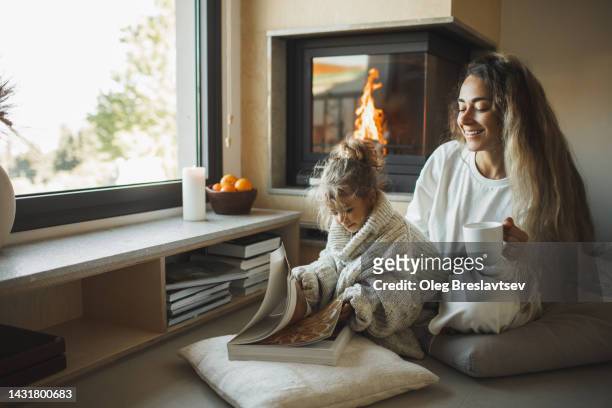 mom and little daughter reading a book together near fireplace at home - cocoon stock pictures, royalty-free photos & images