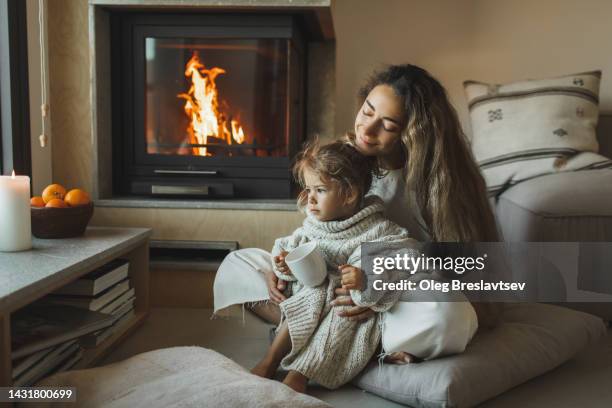 mom with cute daughter enjoying warm cocoa near fireplace. christmas family evening at home. - skandinavische kultur stock-fotos und bilder
