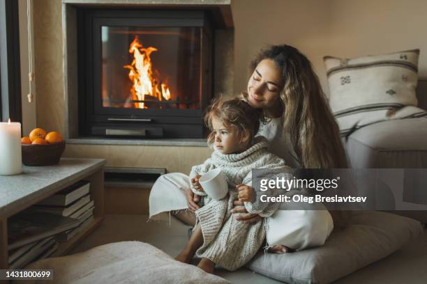 mom with cute daughter enjoying warm cocoa near fireplace. christmas family evening at home. - elegance family stock pictures, royalty-free photos & images