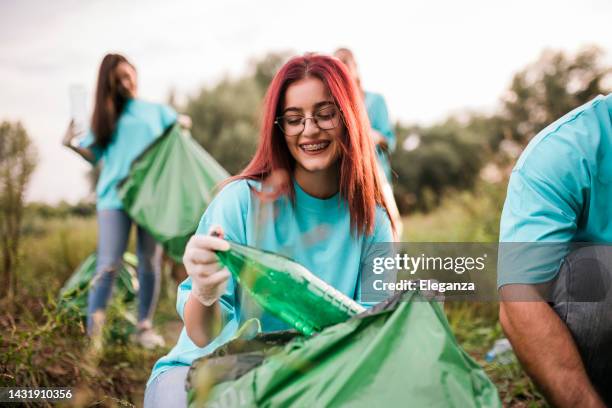 grupo de voluntários usando luvas de proteção coletando lixo em uma reserva natural por uma floresta durante um dia de limpeza. na frente está uma ruiva ruiva bonita de pé com os braços cruzados, todos usando um top azul, sentindo-se bem pelo que ele - responsabilidade social - fotografias e filmes do acervo