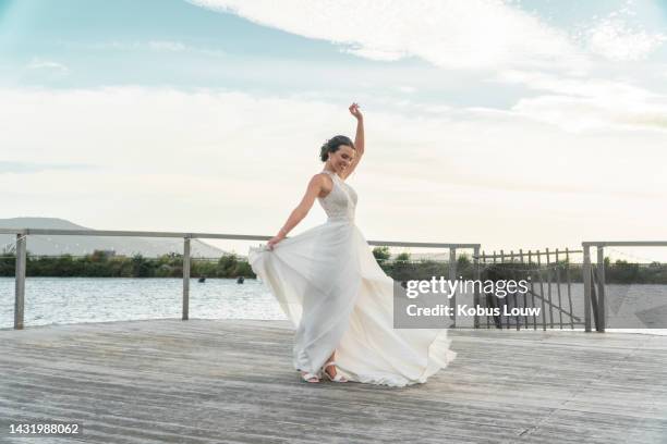 bride with white wedding dress, happy to pose outdoor on a jetty with lake or dam in background. woman smile on day of marriage celebration outside with water, trees and mountain in nature backdrop - bride walking stock pictures, royalty-free photos & images