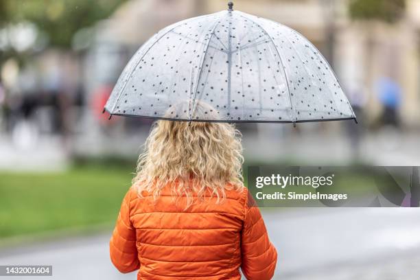 woman in orange jacket walks in the city during a rainy day holding a seethrough umbrella above her. - spring weather stock pictures, royalty-free photos & images