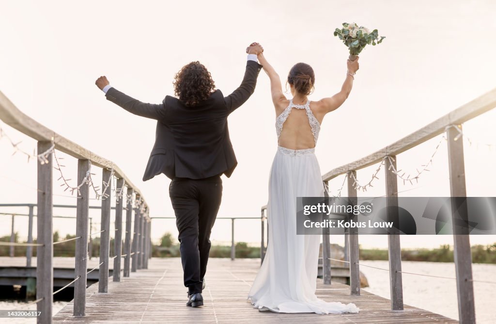Couple, walking and wedding, celebration of love with hands in sky and bridge at lake or dam together. Man, bride with bouquet of flowers and walk by river, happy on day of marriage in summer
