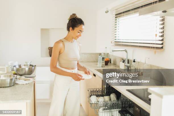 young woman loads  plates and cups in the dishwasher. household chores with pleasure. - woman cleaning for man stock-fotos und bilder