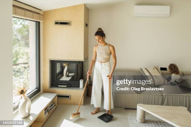 young woman sweeping the floor and cleaning house while baby watching cartoons on mobile phone - agent de ménage photos et images de collection