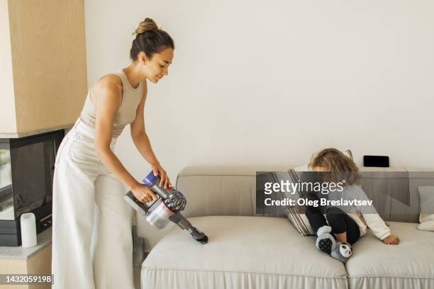 smiling mother showing to daughter how to vacuum dust on sofa with small portable vacuum cleaner - vacuum cleaner woman stockfoto's en -beelden