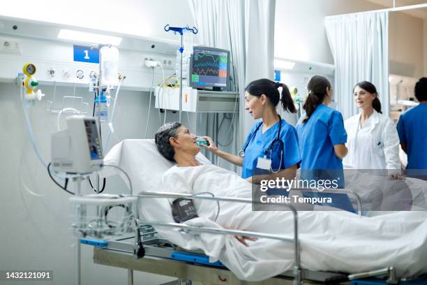 nurse adjusting medical equipment while speaking with female patient in open plan hospital ward with patients and medical personnel - general view stock pictures, royalty-free photos & images