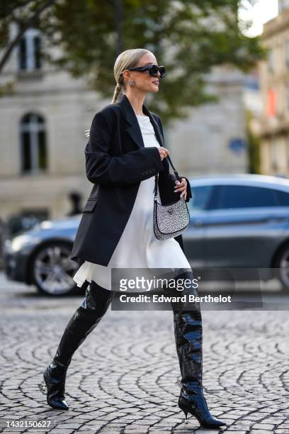Leonie Hanne wears black sunglasses from Loewe, diamonds logo earrings from Balanciaga, a black blazer jacket, a white shoulder-off / knees dress, a...