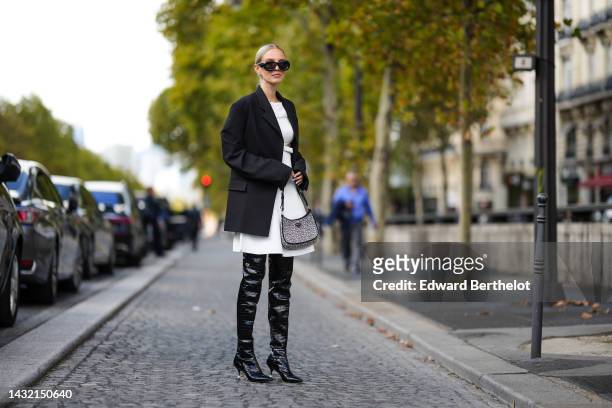 Leonie Hanne wears black sunglasses from Loewe, diamonds logo earrings from Balanciaga, a black blazer jacket, a white shoulder-off / knees dress, a...
