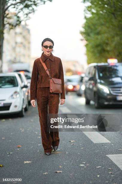 Mary Leest wears black sunglasses, gold earrings, a brown turtleneck pullover, a dark brown embossed velvet ribbed shirt, matching dark brown...