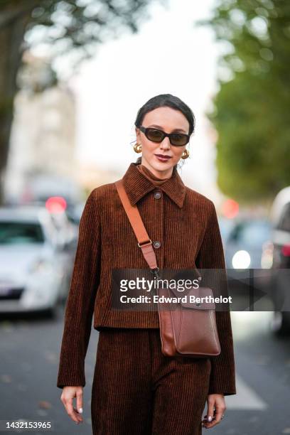Mary Leest wears black sunglasses, gold earrings, a brown turtleneck pullover, a dark brown embossed velvet ribbed shirt, matching dark brown...